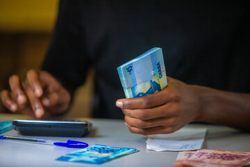 vertical shot of Dark skinned African person working on desk holding counting Ghanaian cedi notes money with some notes on table. accounting concept. 