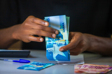 vertical shot of Dark skinned African person working on desk holding counting Ghanaian cedi notes money with some notes on table. accounting concept. 