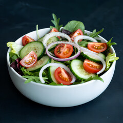 Close-up of a fresh vegetable salad with lettuce, tomatoes, and cucumber in a white bowl on a dark background.