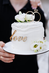 Elegant wedding cake with rings and white flowers, held by newlyweds in celebration