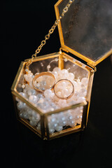 Two gold wedding rings in a glass box filled with decorative pearls on black background