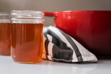 Homemade ghee process in bright natural light, golden clarified butter in glass mason jars beside a red cast iron pot and striped tea towel, high contrast kitchen scene with warm shadows and bright