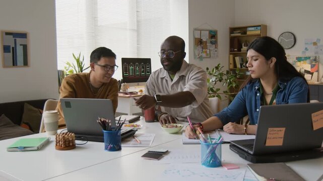 Three biracial developers gathering at desk with laptops and documents, they discussing new application and enjoying fresh baked cookies