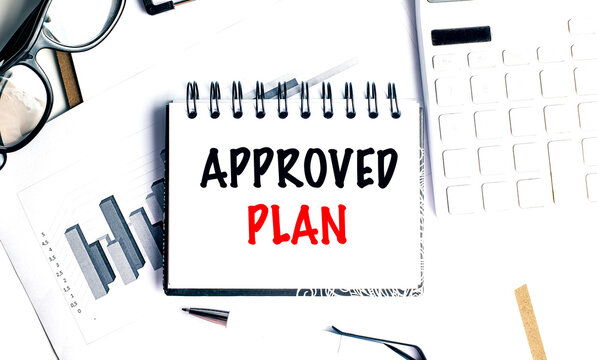 Notebook with text approved plan on white paper surrounded by charts, calculator, pen and glasses on desk
