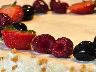 A close-up shot of a delicious-looking cake topped with fresh strawberries, raspberries, and blueberries. The side of the cake is covered in white chocolate flakes.