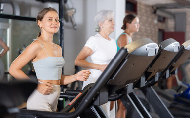 Fototapeta premium Girl walk on treadmill in gym. Active hobby, healthy lifestyle, extensive training. Happy sporty people interacting in weight room training - social gathering concept in sport lifestyle context