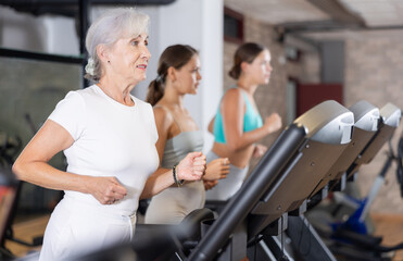 Mature positive woman running on treadmill in modern fitness club