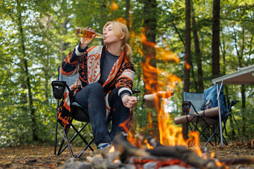 Woman drinking beer while grilling sausages on campfire in forest