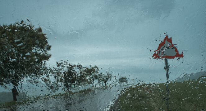 Raindrops on a car windshield distort the view of blurred traffic and green trees, creating a moody, atmospheric perspective of a rainy day drive, selective focus, blurring