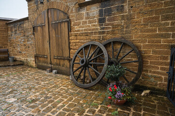 Obraz premium Rustic charm in a vintage courtyard with wooden cart wheels leaning against an old stone wall, cobblestone path, and a vibrant flower pot. Perfect for travel, history, and garden themes