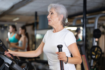 Slender athletic mature woman during cardio exercises on elliptical simulator in fitness center....