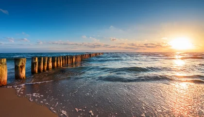 Poster de jardin  Wooden Breakwater Stretches Into Blue Sea Under Soft Evening Light Gentle Waves Touch Sandy Shore While Sun Reflects Warmly On Weathered Timber Posts And Shimmering Water  © Tatianna