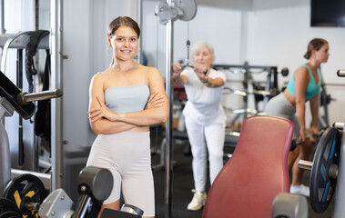 Fototapeta premium Young girl trainer in sportswear poses in middle of fitness center gym. Active lifestyle, multi-age group, daily workouts in crossfit gym