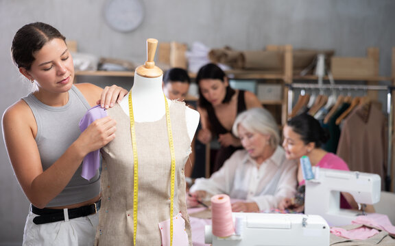 Young female fashion design student shaping garment on mannequin using pins during practical workshop, surrounded by classmates engaged in assembling fabric clothing parts on sewing machines - Powered by Adobe