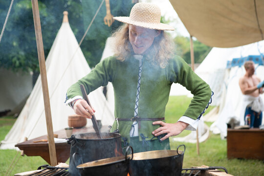 Smiling young Man in medieval attire straw hat and green tunic, cooks over open flame at historical reenactment event. Reconstructions provide valuable insights into daily life, past eras traditions.