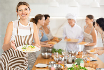 Young woman holding plate with cooked chicken breast in her hands posing surrounded by other members of cooking course.