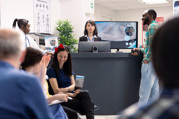 Clinic front desk worker stands at reception, updating medical records on computer with information from doctor. Female hospital receptionist looks at camera, ready to greet and help incoming patients © DC Studio
