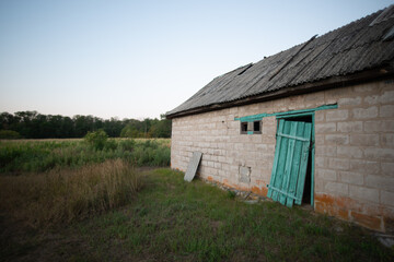 A wide view of an old, abandoned brick outbuilding with a partially collapsed roof, featuring a vibrant turquoise door standing ajar, with a field of dry grass nearby.
