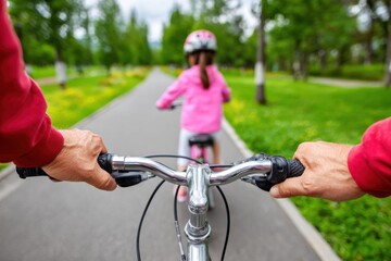 Obraz premium First-person perspective of a parent riding a bicycle behind their child, teaching them to ride in a park on a bright sunny day, creating precious family moments.