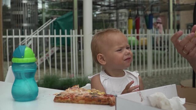 A little girl with short blond hair bites a slice of pizza, gives it to her mother, and takes another huge slice from a table on a cafe's summer terrace on a hot summer day.