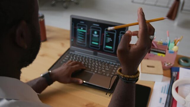 Over shoulder view of unrecognizable black man working on laptop creating app presentation and twirling pencil in hand