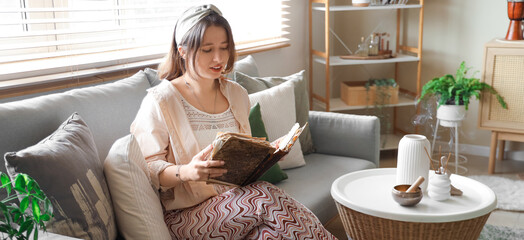 Young witch with spell book sitting on sofa at home