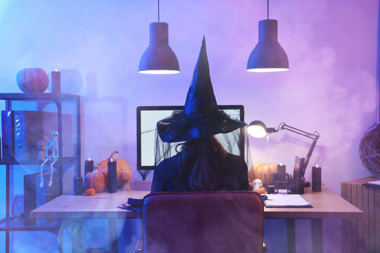 Woman dressed for Halloween as witch using computer at table in dark office, back view
