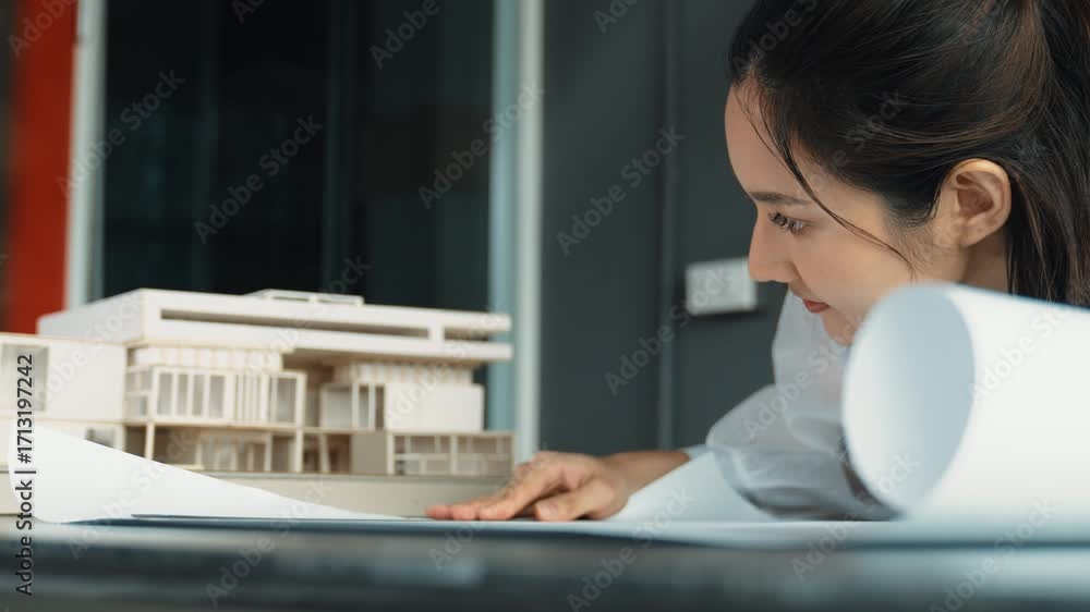 Poster closeup of asian young beautiful architect engineer looking at project plan with model placed on tab