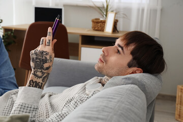 Young man with disposable electronic cigarette resting on sofa at home
