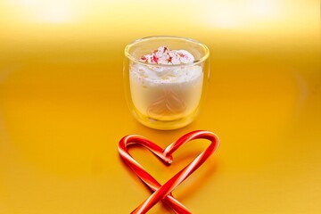 Glass of eggnog with whipped cream, cinnamon and red sugar on top. There are peppermint sticks forming a heart in the foreground. The background is metallic gold.