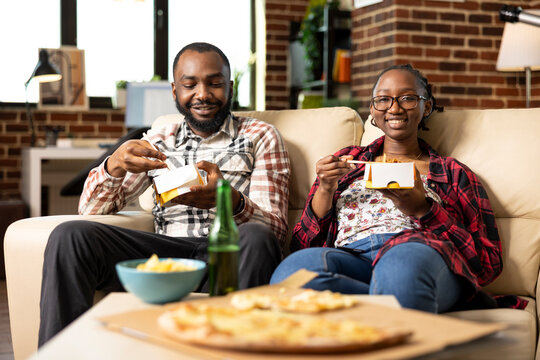 Happy male and female friends seated on couch holding takeout boxes, having lunch at home. African american couple having cozy weekend hangout with food and drinks in brick wall apartment. - Powered by Adobe
