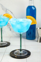 Glasses of tasty Blue Lagoon cocktail garnished with orange on white tile table