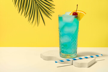Glass of tasty Blue Lagoon cocktail garnished with cherry and palm leaf on white table against yellow background