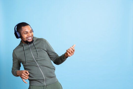 African american upbeat man pretending to play air guitar and listening to music on his headset, showing a good mood with his rock and roll attitude. Lively male dancer against blue background. - Powered by Adobe