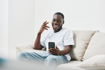 Happy African American man enjoying a video call on his smartphone while relaxing on a sofa, wearing casual clothing in a bright living room