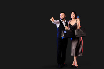 Young couple with credit card and shopping bags pointing at something on dark background. Cyber Monday