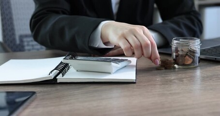 Woman putting coins into glass jar and using calculator at wooden table indoors, closeup - Powered by Adobe