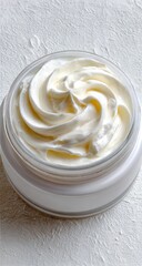 A close-up reveals a creamy, off-white substance swirled in a clear, round jar atop a textured white background.  The substance is thick and appears smooth, suggesting a rich texture