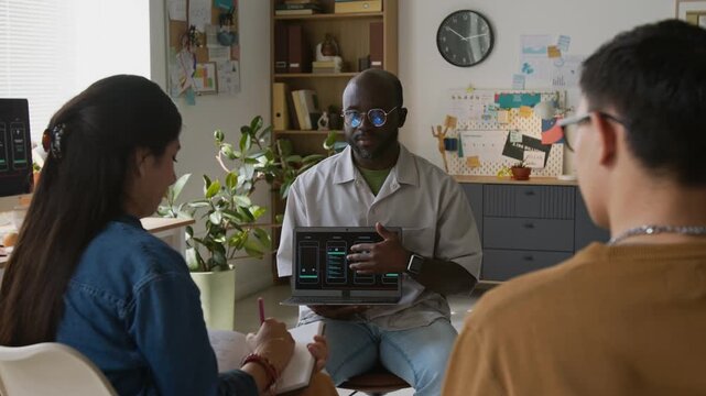 African American teamlead sitting in office chair in front of two Caucasian programmers and presenting new project