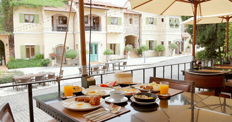 Mediterranean breakfast on outdoor cafe terrace, village architecture background, sunny morning. Fresh orange juice, eggs, pastries, relaxing ambiance, European dining experience.