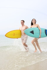 Obraz premium Couple lover running on beach with surfboards, splashing water and smiling under summer sunlight, concept of friendship, vacation, holidays, surfing, freedom, and fun tropical lifestyle. 
