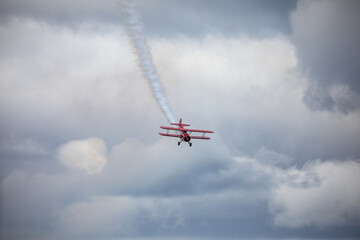 A restored World War II era red biplane airplane performing aerobatic maneuvers in the sky during an air show event, leaving a trail of smoke against a cloudy background in Hillsboro, Oregon, USA