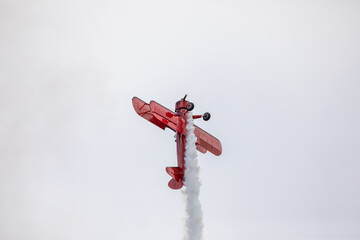 A restored World War II era red biplane airplane performing aerobatic maneuvers in the sky during an air show event, leaving a trail of smoke against a cloudy background in Hillsboro, Oregon, USA