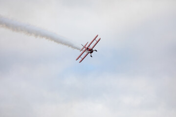 A restored World War II era red biplane airplane performing aerobatic maneuvers in the sky during an air show event, leaving a trail of smoke against a cloudy background in Hillsboro, Oregon, USA