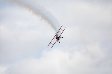 A restored World War II era red biplane airplane performing aerobatic maneuvers in the sky during an air show event, leaving a trail of smoke against a cloudy background in Hillsboro, Oregon, USA