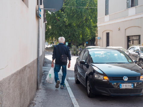 Senior man walking along an urban sidewalk after shopping, carrying a reusable grocery bag, with a black Volkswagen Polo V 6R hatchback parked on the street