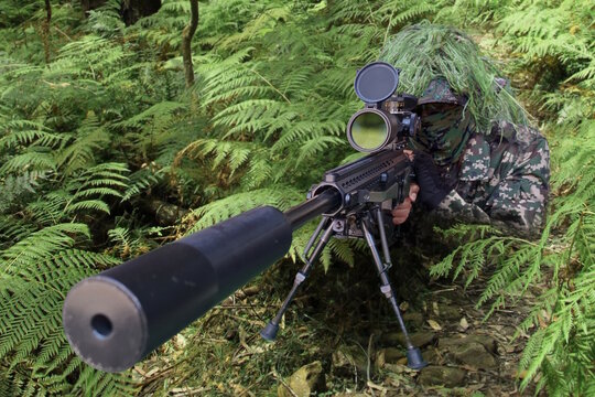 Military sniper with camouflage ghillie suit, positioned in a fern-covered area aiming with a precision rifle. 