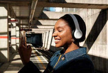 Young black woman listening music with headphones and using smartphone in subway station