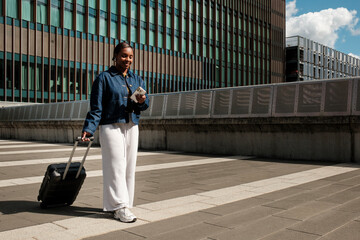 Fototapeta premium Young black woman using smartphone while pulling luggage in city