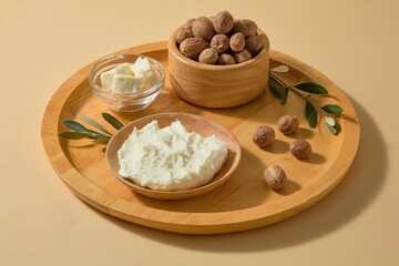 Wooden spoon scooping creamy shea butter beside loose nuts on a neutral white table, perfect for highlighting natural cosmetic ingredients or DIY beauty visuals.
